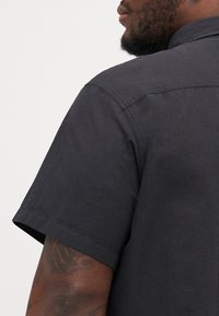 Chemise noire à manches courtes avec un col structuré, présentant une texture lisse et des coutures d'épaule renforcées. Vue incomplète montrant le tombé du tissu.