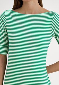 Nő zöld-fehér vízszintesen csíkos, testhezálló felsőt visel, rövid ujjakkal és széles csónaknyakú kivágással.