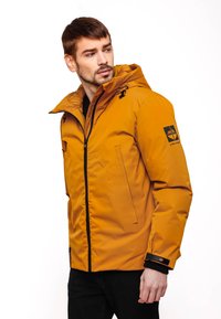 Giacca impermeabile gialla con zip frontale, cappuccio, tasche laterali e dettagli neri sui polsini. Presenta un logo sul braccio sinistro.