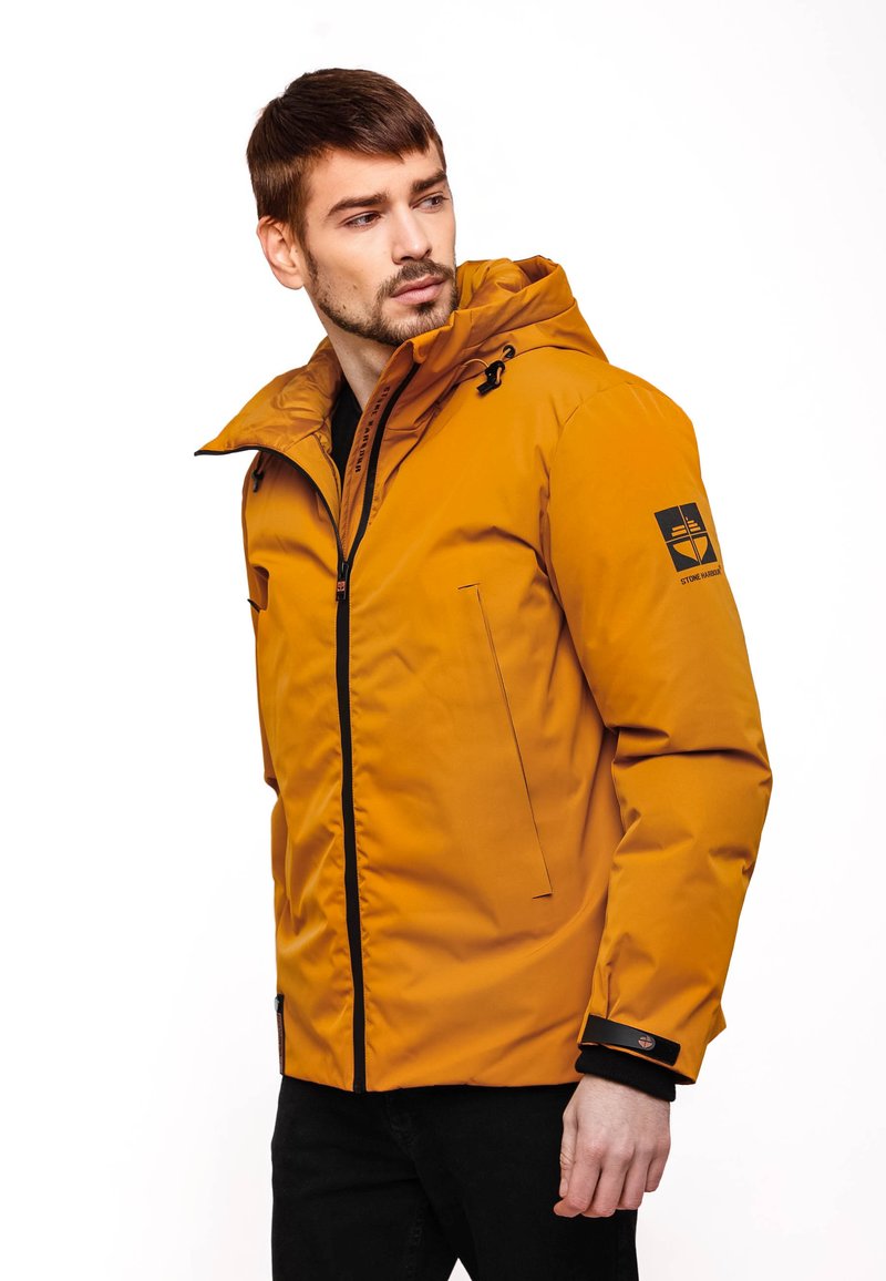 Giacca impermeabile gialla con zip frontale, cappuccio, tasche laterali e dettagli neri sui polsini. Presenta un logo sul braccio sinistro.