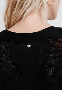 Blondyna tyłem w czarnej dzianinowej bluzce z trójkątną metalową ozdobą przy dekolcie i złotą okrągłą chwilą w uchu.