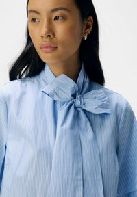 Camicia a righe blu chiaro con un grande fiocco al colletto, realizzata in un tessuto morbido, con sottili righe gialle per creare un contrasto.