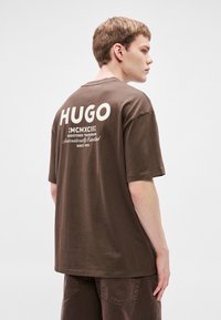 Brązowy bawełniany t-shirt z dużym białym napisem „HUGO” na plecach, krótkimi rękawami i luźnym krojem z prostym dołem.