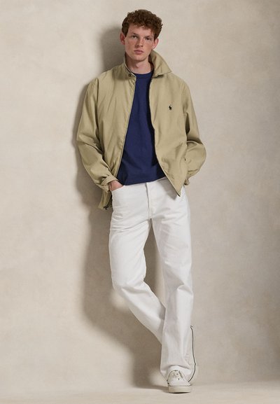 Νέος άνδρας με μπεζ μπουφάν, μπλε σκούρο πουκάμισο, λευκό παντελόνι και λευκά αθλητικά παπούτσια, στέκεται μπροστά σε απλή μπεζ τοίχο με τα χέρια στις τσέπες.