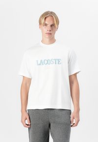 Biela bavlnená tričko s farebným logom "LACOSTE" v tučnom, iridescentnom písmene cez hrudník. Okrúhly výstrih, krátke rukávy.
