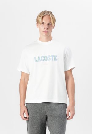Weißes Baumwoll-T-Shirt mit einem mehrfarbigen "LACOSTE"-Logo in fetter, schillernder Schrift über der Brust. Rundhalsausschnitt, kurze Ärmel.
