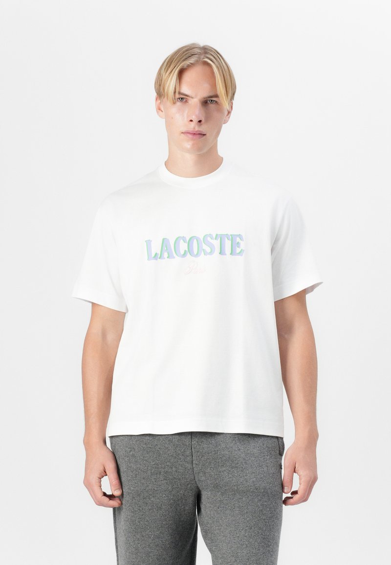 Biela bavlnená tričko s farebným logom "LACOSTE" v tučnom, iridescentnom písmene cez hrudník. Okrúhly výstrih, krátke rukávy.