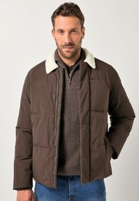 Man in een bruine pufferjack met een witte fleece kraag over een bruine gebreide trui en blauwe jeans, staand tegen een effen achtergrond.