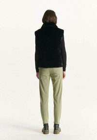 Chaleco de piel sintética negra con cuello alto, combinado con pantalones verde claro. Corte slim, textura suave y diseño sencillo.