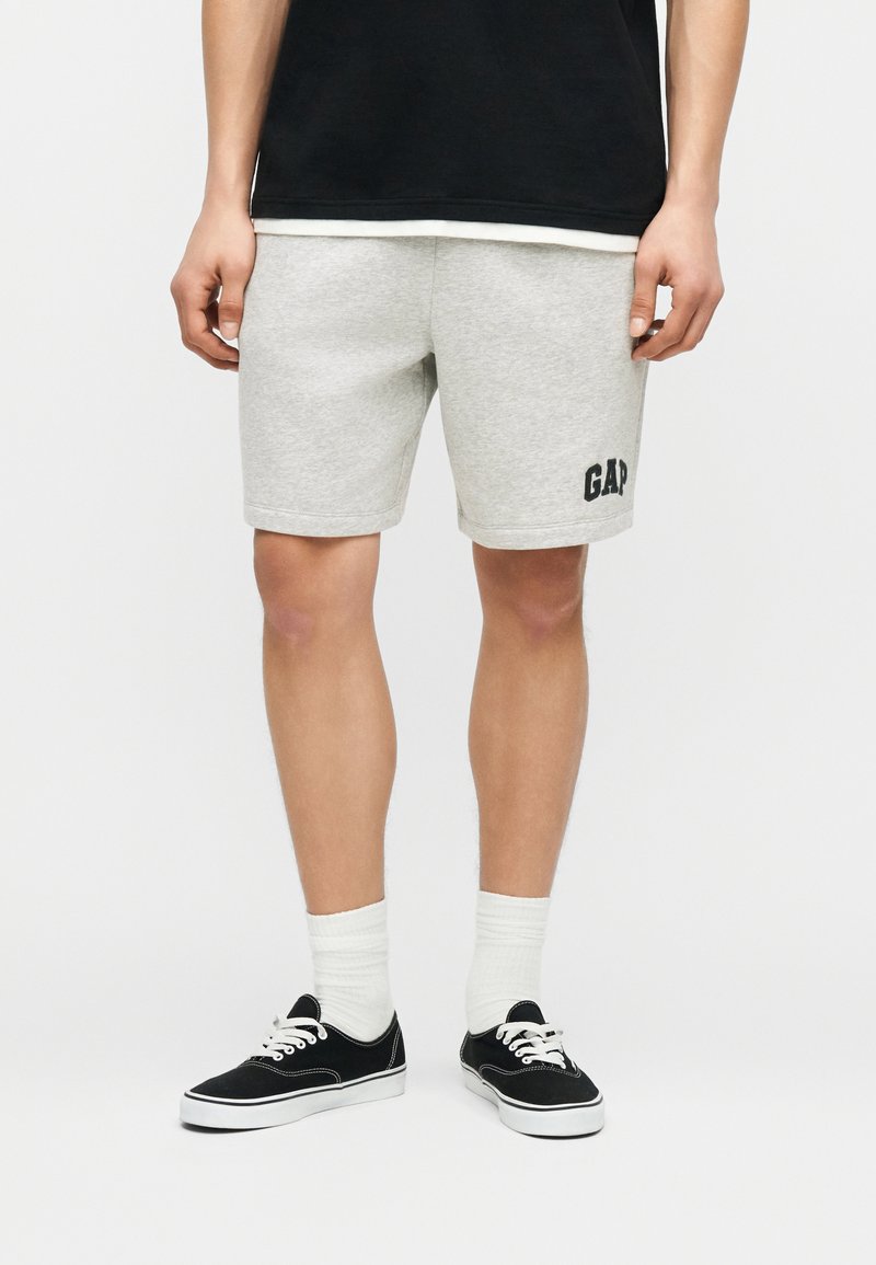 Homme portant un short gris clair avec le logo "GAP" sur la jambe gauche, des chaussettes blanches, des baskets noires et un t-shirt noir, debout devant un fond uni.
