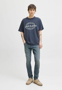 Mörkblå oversized t-shirt med vit grafisk tryck, tillsammans med ljusblå jeans och beige skor. Modellen visar tatuerade armar.