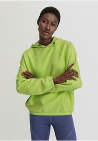 Limonengrüner Fleece-Hoodie mit einem Kordelzugkragen und Raglanärmeln. Bequeme Passform und gerippte Handgelenkbündchen. Getragen mit blauen Sportleggings.