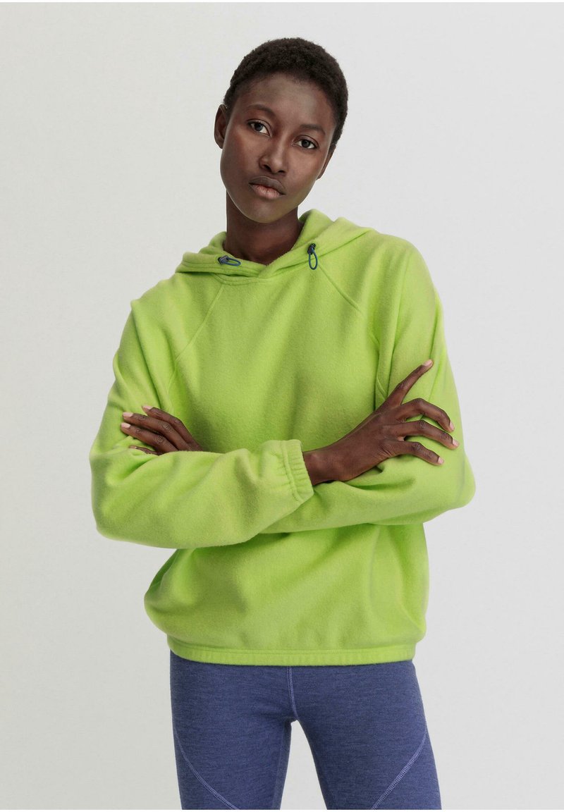 Limonengrüner Fleece-Hoodie mit einem Kordelzugkragen und Raglanärmeln. Bequeme Passform und gerippte Handgelenkbündchen. Getragen mit blauen Sportleggings.