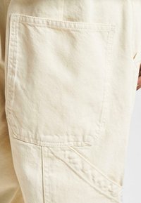 Beige Baumwollhose mit einer großen aufgesetzten Tasche, diagonalen Nähtdetails und einer weichen, strukturierten Oberfläche.
