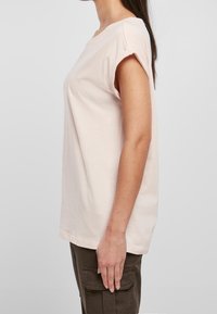 Kobieta w jasnoróżowej koszulce z krótkim rękawem i ciemnych spodniach cargo, ukazana z boku na jednolitym jasnym tle.