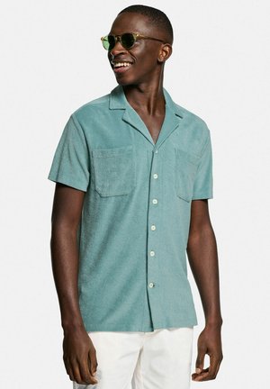 Mintgrünes kurzärmliges Hemd aus strukturiertem Stoff, ausgestattet mit zwei Brusttaschen und weißen Knöpfen, getragen mit einer dunklen Sonnenbrille.