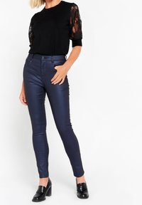 Mustat yläosa pitsihihoilla, yhdistettynä tummansinisiin kiiltäviin slim-fit-housuihin ja mustiin korkokenkäloafereihin. Suora siluetti ja sileät tekstuurit.