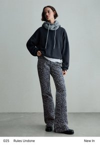 Čierna oversized mikina s šedým fleecom na golieri, kombinovaná s rozšírenými, vzorovanými nohavicami v tmavých odtieňoch. Hladká textúra, pod ňou vrstvená biela košeľa.