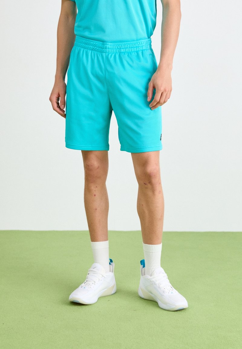 Personne portant un short de sport bleu vif, des chaussettes hautes blanches et des baskets blanches, debout sur un sol vert avec un fond uni.