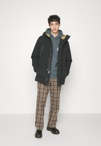 Black parka jacket with a beige lining, front pockets, paired with a blue hoodie and brown plaid trousers, worn with black boots.