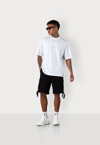 White cotton t-shirt with logo at chest, paired with black cargo shorts. Model wears sunglasses and white sneakers with textured accents.