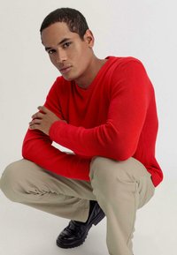 Roter Strickpullover mit V-Ausschnitt und slim-fit Ärmeln; kombiniert mit hellbeigen Hosen und schwarzen Schuhen. Subjekt hockt, Arme verschränkt.