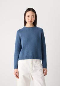 Blauwe gebreide trui met een textuurafwerking, ronde halslijn en geribbelde zoom. Gecombineerd met witte broek, die een relaxte pasvorm laat zien.