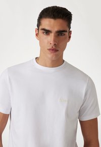 Camiseta blanca de algodón con mangas cortas y cuello redondo. Presenta un logo sutil bordado en beige claro en el lado izquierdo del pecho.