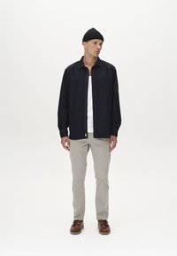 Dunkelblaue Jeanshemd, langärmlig, mit Knopfleiste; darunter weißes Top. Hellgraue Hose und braune Schuhe runden das Outfit ab.