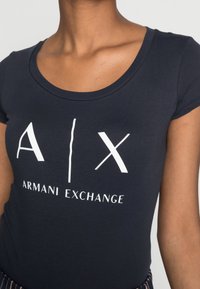 Close-up de uma pessoa a usar uma t-shirt Armani Exchange azul-marinho com o logótipo e texto brancos "A | X" no peito.