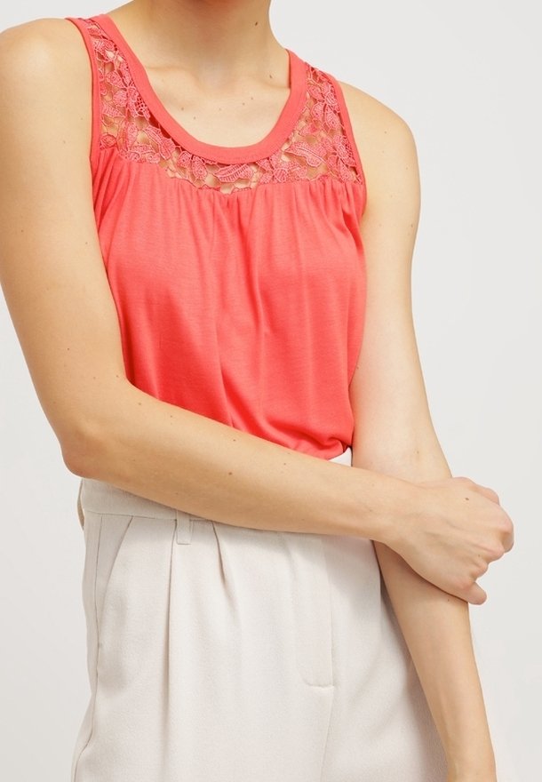 Femme portant un haut sans manches corail avec un détail en dentelle et un pantalon blanc taille haute, tenant son bras gauche avec sa main droite.