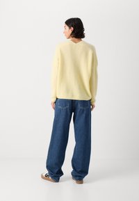 Gelber Strickpullover mit lockerer Passform, V-Ausschnitt und überschnittenen Schultern, kombiniert mit blauen Denim-Jeans im Weitschnitt.
