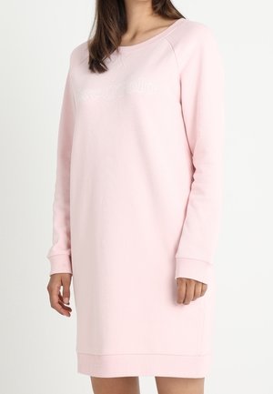 Kobieta w jasnoróżowej, długorękawowej sukience typu sweatshirt z delikatnym wypukłym napisem na piersi, stojąca na jednolitym białym tle.