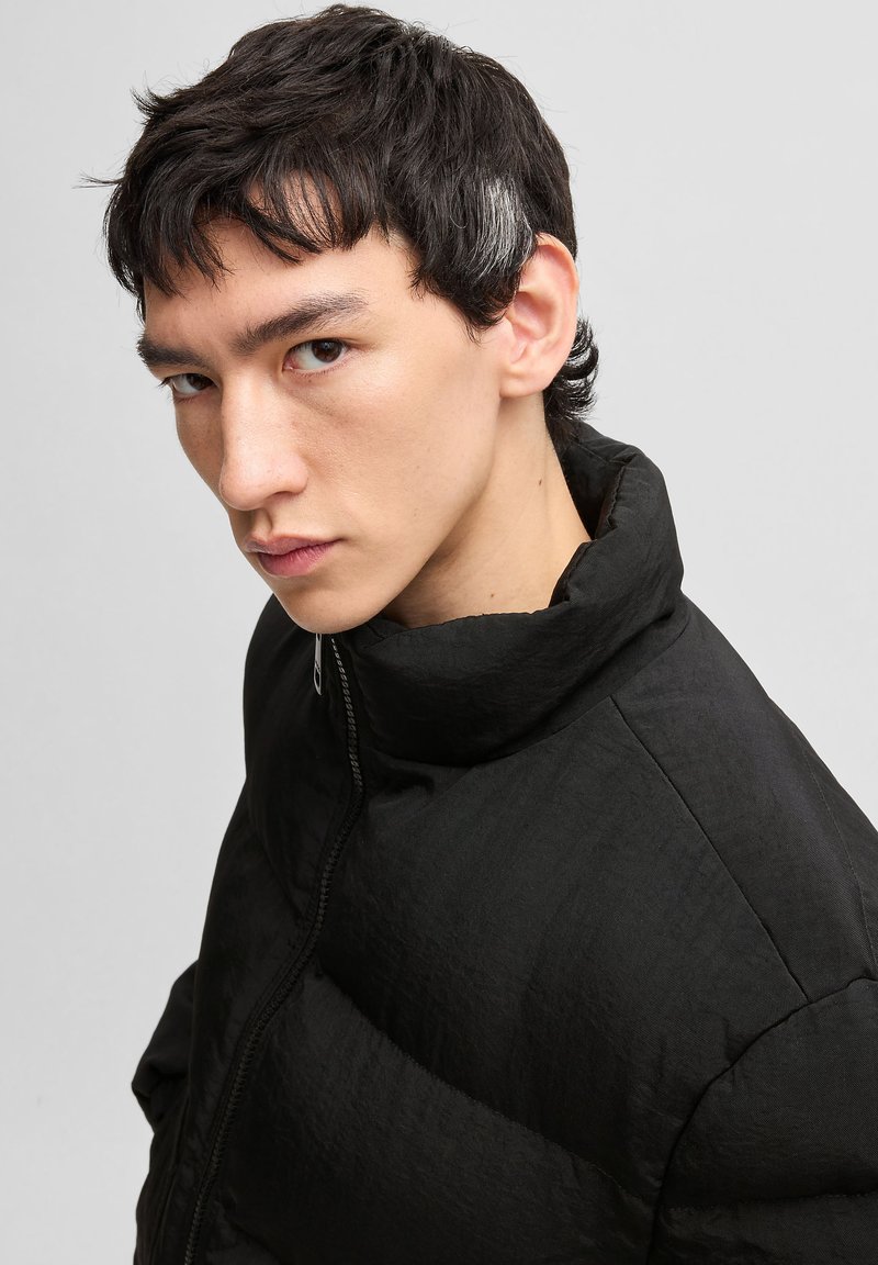 Doudoune noire avec un col haut et une fermeture éclair. Présente un tissu texturé et un design matelassé pour l'isolation et la chaleur.