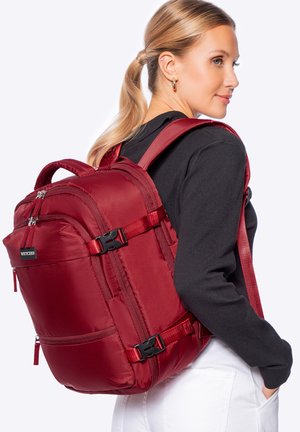 Blonde vrouw in een zwarte top en witte broek met een grote rode rugzak met meerdere ritsen en verstelbare banden.