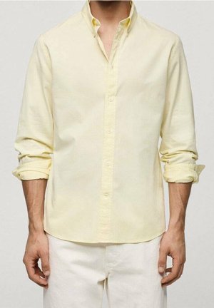 Mężczyzna ubrany w bladoróżową, długorękawową koszulę z podwiniętymi rękawami i białe spodnie na jednolitym tle.