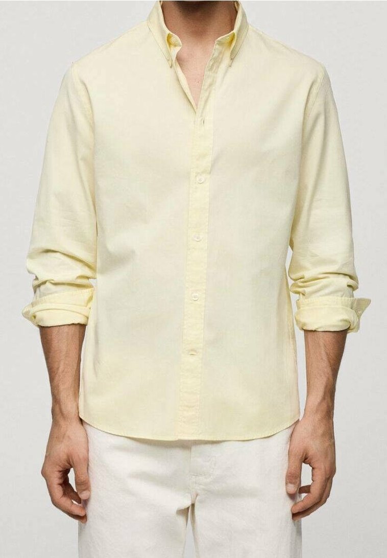 Homme portant une chemise jaune pâle à manches longues, boutonnée, avec les manches retroussées et un pantalon blanc sur un fond uni.