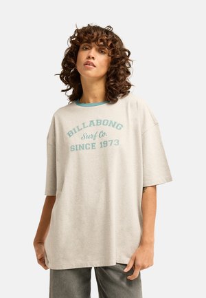 Femme aux cheveux bouclés portant un t-shirt Billabong beige clair oversize avec un texte bleu et un pantalon foncé, debout devant un fond uni.