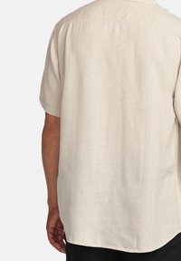 Camisa de botones de manga corta, beige, hecha de tejido texturizado. Presenta un corte relajado con un dobladillo recto y líneas sutiles en el material.
