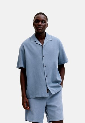 Hombre de piel oscura con camisa de manga corta azul claro abotonada y pantalones cortos a juego, de pie con una mano en el bolsillo contra un fondo liso.