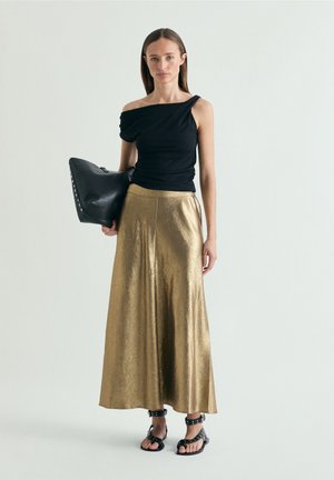 Femme portant un top asymétrique noir, une jupe métallisée dorée longueur cheville, des sandales noires à clous, tenant une grande pochette en cuir noir.