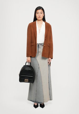Kvinne som har på seg brun blazer, hvit snørtopp, lang to-farget denimskjørt, svarte spisse sko, holder en svart ryggsekkveske mot en ensfarget bakgrunn.