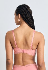 Rosa blonde BH med justerbare stropper, intrikate bølgekantdetaljer, og en krok-lukking bak, designet med en glatt overflate.