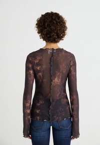 Haut transparent à manches longues avec un motif tie-dye marron et des bords festonnés. Doté d'une fermeture au dos ; associé à un jean bleu ajusté.