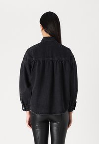Musta denim-takki, jossa on rento istuvuus, napitettu hihanpää ja kaareva helma. Ominaisuuksiin kuuluu möykkykuvio ja pehmeä tekstuuri. Näyttää takaa.