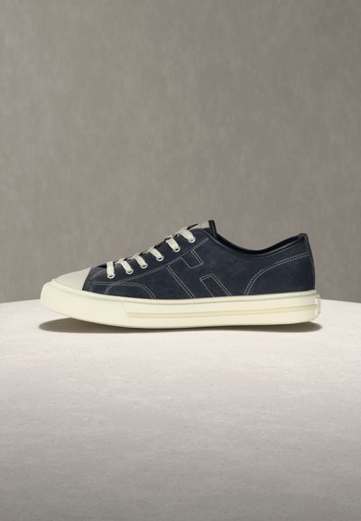 Pantof sport de culoare albastru închis, cu talpă joasă, șireturi albe, capac de cauciuc alb pe vârf și talpă groasă albă, pe o suprafață simplă, cu un fundal gri.