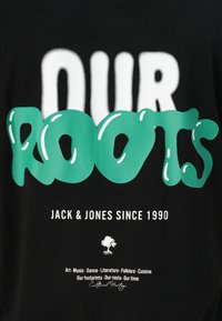 Camiseta negra con una gran tipografía blanca y verde que dice "NUESTRAS RAÍCES" junto a un gráfico de un árbol y texto sobre arte y cultura a continuación.