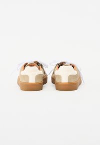 Beiga mockasin-sneakers med vita accenter, brun gummisula, vita snören och en dragkedjedetalj bak. Mjuk textur och rund form.