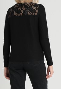 Maglia nera a maniche lunghe con tessuto testurizzato e design in pizzo sulla schiena, caratterizzata da motivi floreali. Abbinata a jeans scuri.