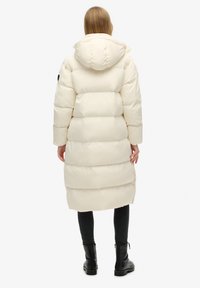 Lang, puffy, cremefarvet jakke med hætte, der har horisontale quiltninger, sidelommer og en afslappet pasform. Sorte accenter på ærmerne.